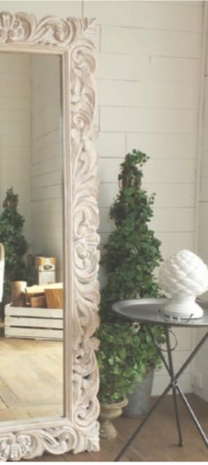 Decorative white mirror with intricate carvings in a room with plants and a table.