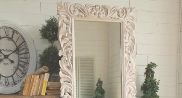 Decorative mirror with intricate carvings on a white wall with a clock and plants.