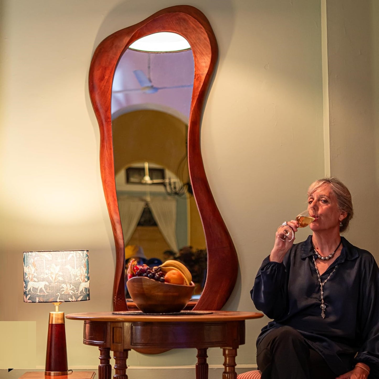 Woman sitting in a room with a unique mirror and a table with fruit and a lamp.