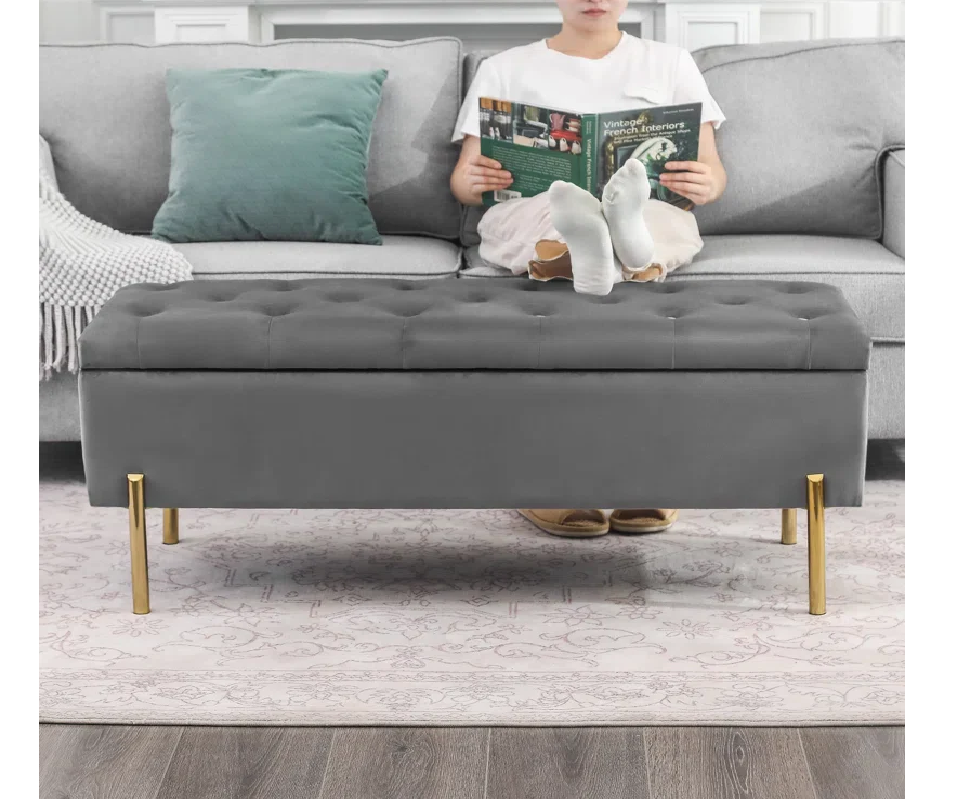 Person sitting on a gray tufted bench in a living room with a couch and pillows.
