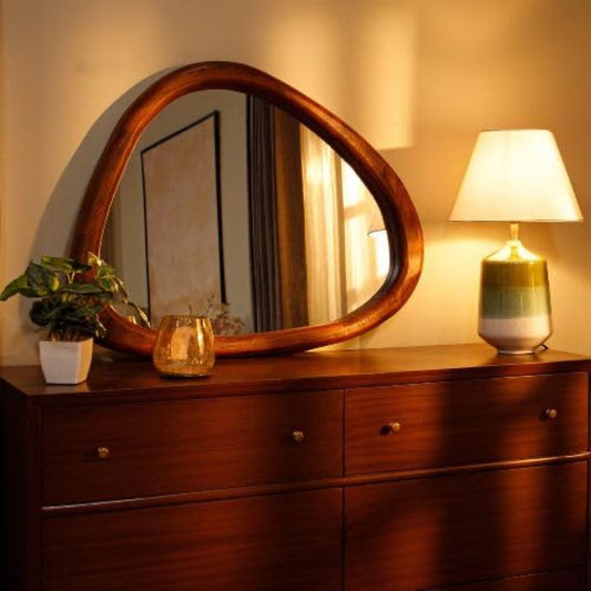 Wooden dresser with round mirror, lamp, and plant in a warm-toned room.