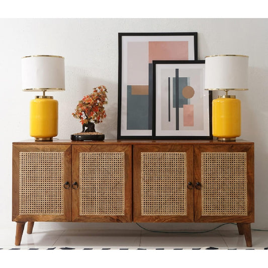 Wooden sideboard with rattan doors, two yellow lamps, a decorative plant, and framed art on a white wall.