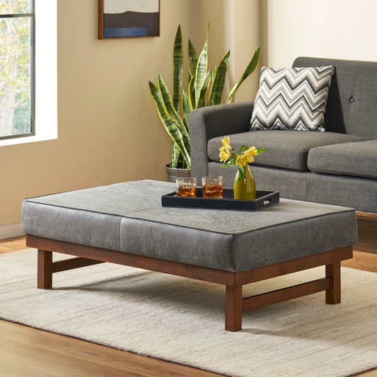 Gray upholstered coffee table with a tray of drinks and flowers in a living room setting.