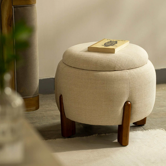 Beige ottoman with wooden legs and a book on top in a room setting.