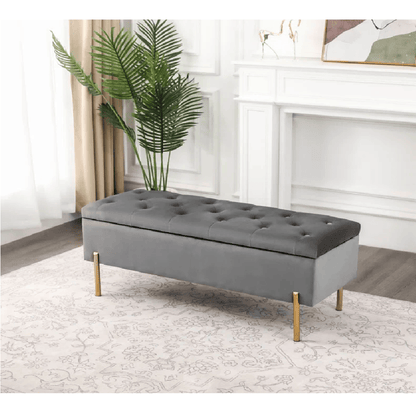 Gray tufted bench with gold legs in a room with a plant and fireplace.