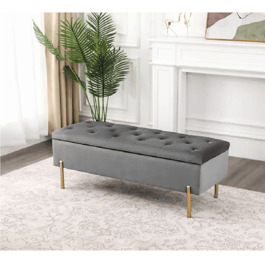 Gray tufted bench with gold legs in a room with a plant and fireplace.