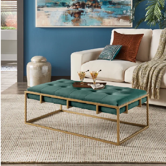 Teal ottoman with gold frame in front of a beige sofa with decorative pillows and a painting on the wall.