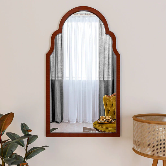 Arched wooden mirror on a white wall with a reflection of a yellow chair and curtains.